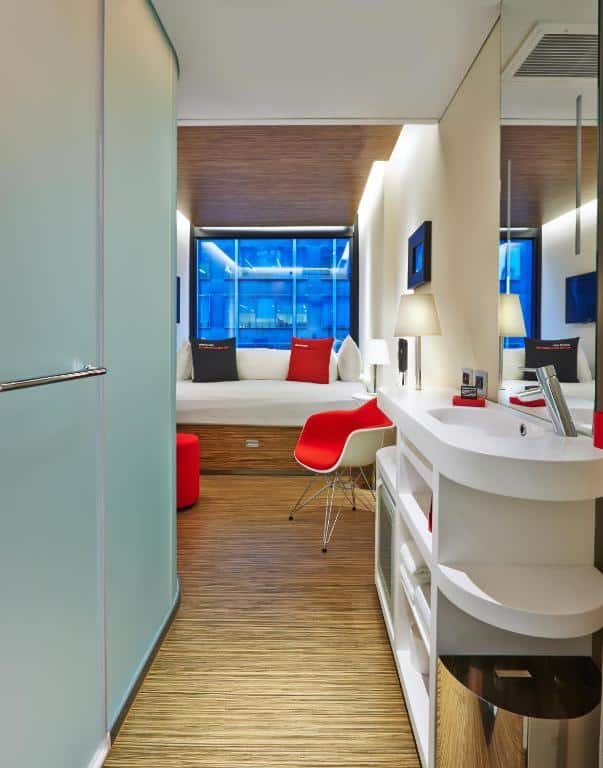 quarto do citizenM New York Times Square mostrando uma cama de casal ao fundo do quarto, de frente para câmera, com uma janela atrás. No lado direito do quarto há uma pia banca com espelho de frente para a porta de vidro do banheiro.