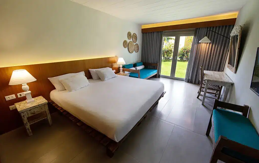 Quarto do Club Med Trancoso com cama de casal ao centro, duas cômodas de madeira em cor branca com luminária ao lado da cama, do lado esquerdo um sofá e em frente uma mesa de trabalho do lado esquerdo portas de vidro com acesso a varanda térrea.