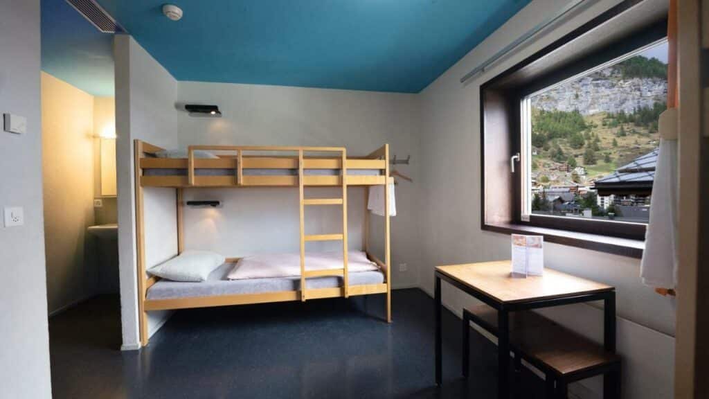 Quarto compartilhado do Zermatt Youth Hostel com uma beliche no centro da imagem ao fundo e do lado direito uma mesa quadrada com dois bancos. Representa onde ficar em Zermatt.