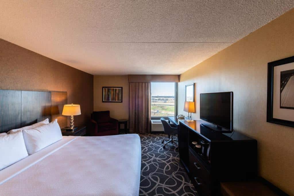 Imagem de um quarto no Crowne Plaza Newark Airport. O quarto possui uma decoração moderna e elegante, com móveis confortáveis e uma cama king-size. À direita, há uma escrivaninha com uma cadeira ergonômica para trabalho ou estudo. Na parede oposta à cama, há uma TV de tela plana. O quarto também inclui um armário para guardar pertences pessoais. A iluminação é suave e agradável, criando uma atmosfera relaxante. O Crowne Plaza Newark Airport é um dos hotéis perto do aeroporto de Newark.