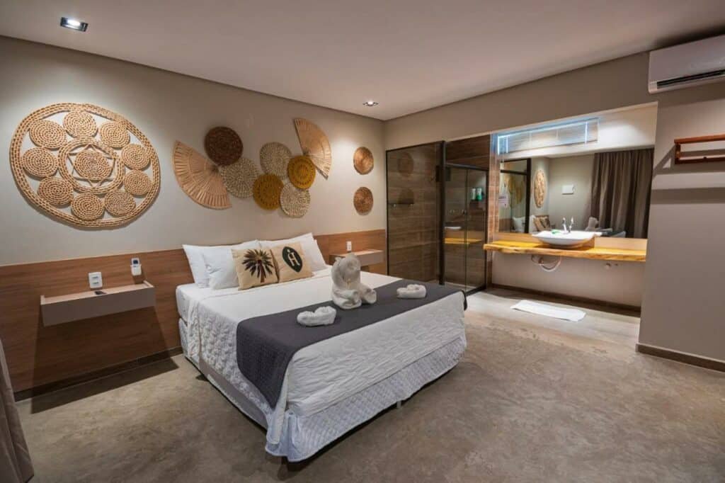 Quarto com cama de casal do Árvo Boutique Hotel, em Maragogi, itens decorativos artesanais acima da cama, ar-condicionado e, à direta, tem um balcão da com pia, espelho e um box de vidro para banho