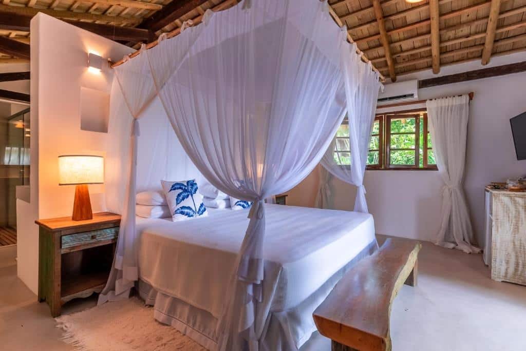 Quarto do Da Graça Hotel com cama de casal ao centro do quarto, com cômoda com luminária do lado direito, um banco de madeira ao pé da cama e do lado esquerdo, uma cômoda de madeira com TV em cima.