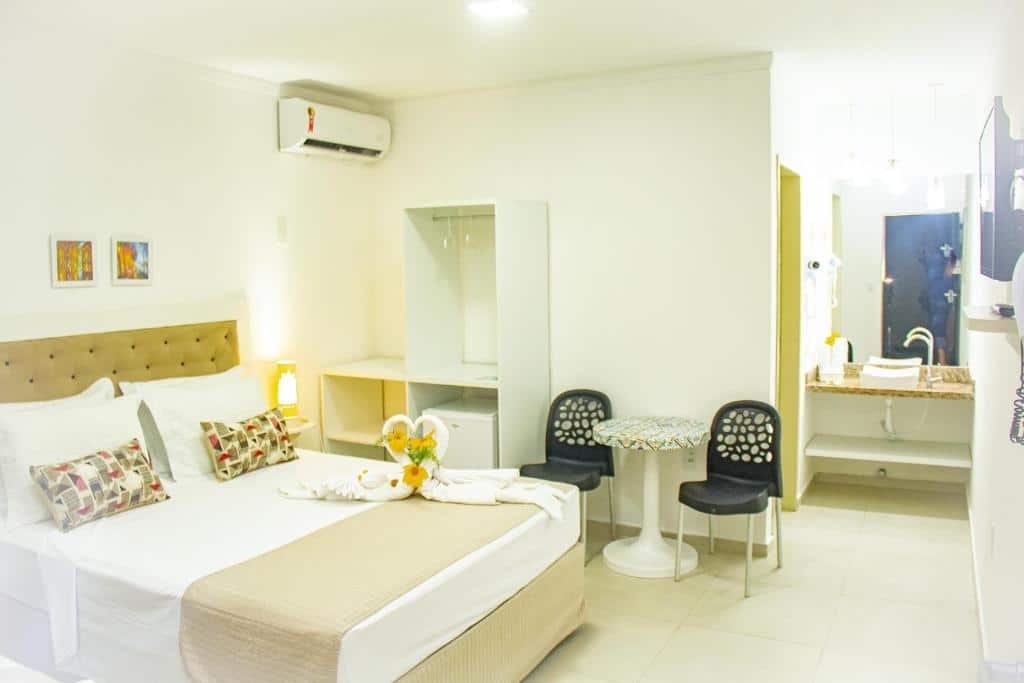 Quarto da Pousada Polymar, de 23 m², com uma cama de casal, ar-condicionado, à direita tem um armário sem portas com repartições para guardar utensílios e um frigobar, ao lado tem uma mesa pequena redonda com duas cadeiras e, mais ao lado, tem uma pia com espelho e uma porta dando acesso ao banheiro