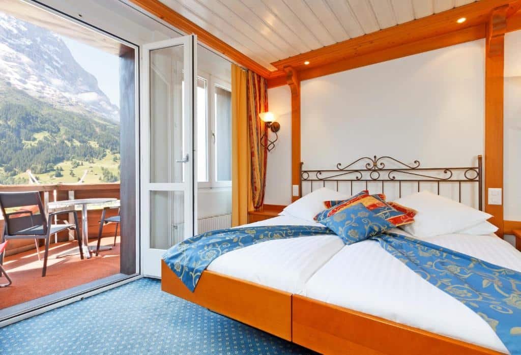 Quarto do Derby Swiss Quality Hotel com cama de casal do lado direito da imagem e do lado esquerdo da cama uma porta que dá acesso a varanda. Representa hotéis em Lauterbrunnen.