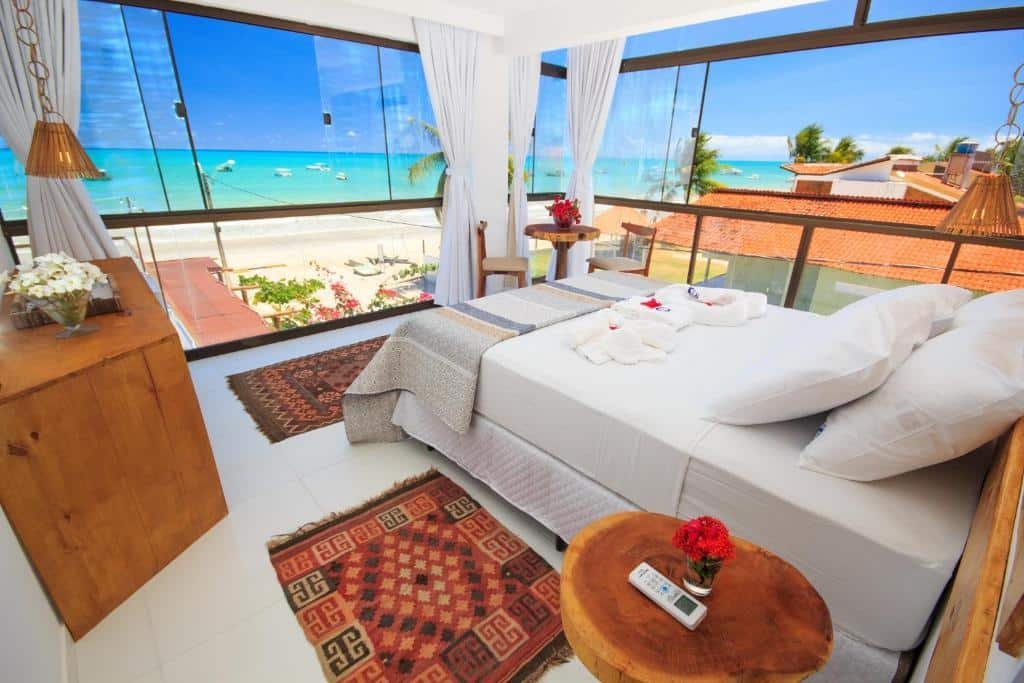 quarto do Ecohar Yoga, com uma cama de casal e paredes de vidro com vista panorâmica do mar azul com alguns barcos navegando
