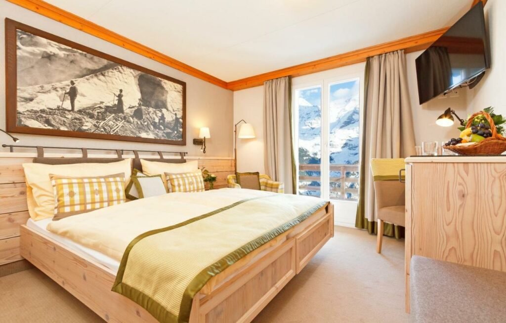 Quarto do Eiger Mürren Swiss Quality Hotel com cama de casal do lado esquerdo da imagem com uma poltrona do lado esquerdo da cama e em frente uma mesa com cadeira e uma TV presa na parede. Representa hotéis em Lauterbrunnen.
