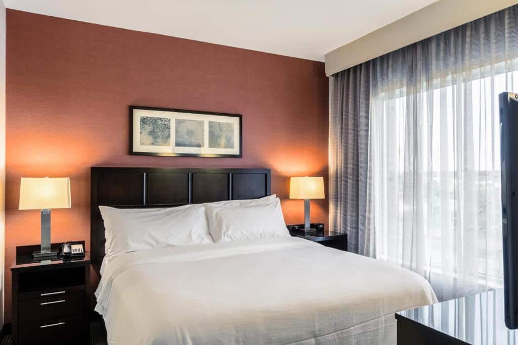 Imagem de um quarto no Embassy Suites by Hilton Newark Airport. No centro do quarto, há uma cama king-size com lençóis brancos e travesseiros macios. À direita da cama, há uma mesa de cabeceira com uma luminária de leitura. Na parede à esquerda da cama, há uma TV de tela plana montada. Ao lado direito da imagem, encontra-se uma área de estar com um sofá confortável e uma mesa de centro. Próximo à janela, há uma escrivaninha com uma cadeira. No canto oposto do quarto, há um armário espaçoso para guardar pertences pessoais. O quarto é iluminado de forma suave e possui uma atmosfera aconchegante. O Embassy Suites by Hilton Newark Airport é um dos hotéis perto do aeroporto de Newark.