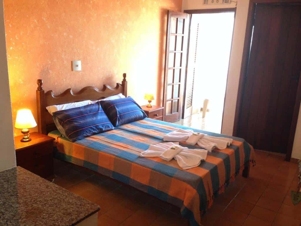 Quarto no Enseada Hotel, uma cama de casal, com toalhas e travesseiros sob a cama, uma sacada, um banheiro e um abajur ao lado da cama, para representar pousadas na Praia da Enseada em Ubatuba