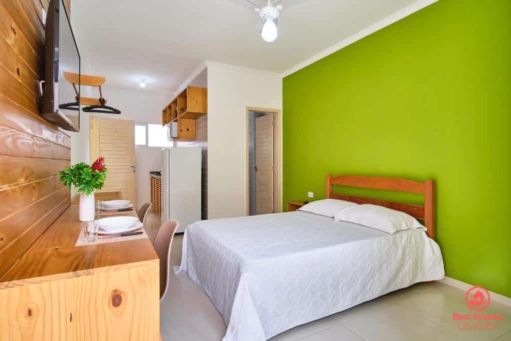 Quarto em Estúdios 3 Praias, um local amplo com uma cama de casal, uma mesa com duas cadeiras, uma televisão, uma geladeira e alguns móveis, para presentar pousadas na Praia da Enseada em Ubatuba