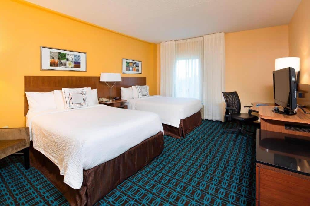 Imagem de um quarto no Fairfield Inn & Suites. No centro do quarto, há uma cama king-size com lençóis brancos e travesseiros macios. À direita da cama, há uma mesa de cabeceira com uma luminária de leitura. Na parede à esquerda da cama, há uma TV de tela plana montada. Ao lado direito da imagem, encontra-se uma área de trabalho com uma escrivaninha e uma cadeira. No canto oposto do quarto, há um armário embutido para guardar pertences pessoais. O quarto é iluminado de forma suave e possui uma atmosfera confortável. O Fairfield Inn & Suites é um dos hotéis perto do aeroporto de Newark.