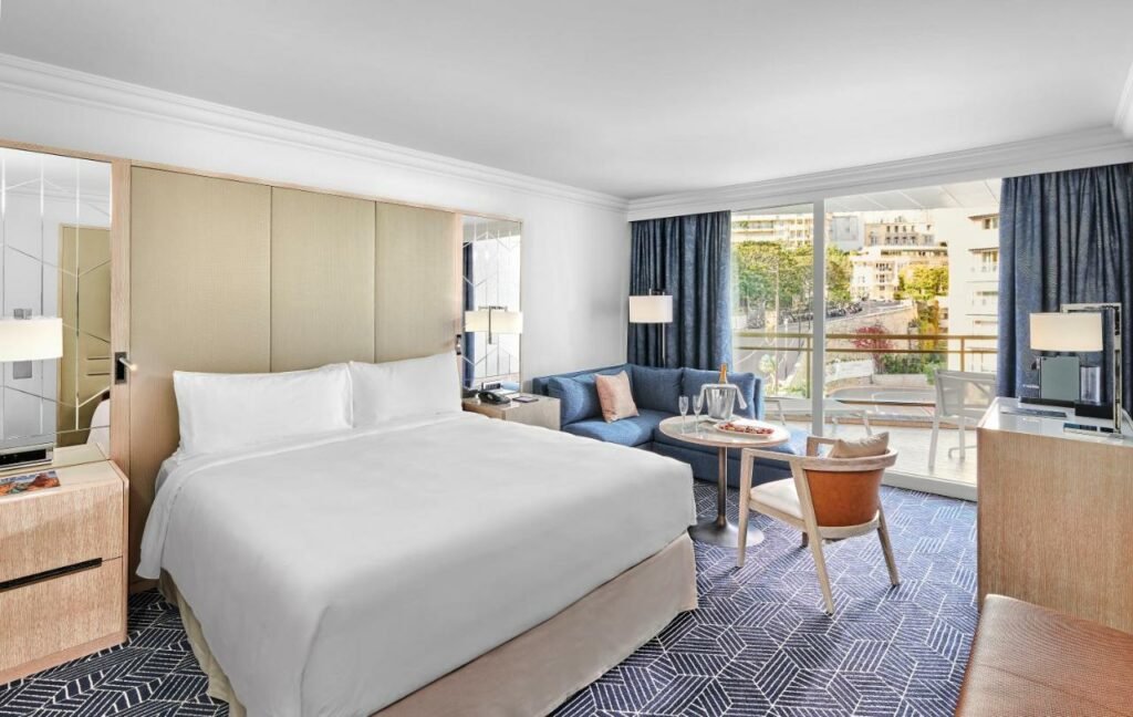 Quarto do Fairmont Monte Carlo. Uma cama de casal no lado esquerdo. Do lado esquerdo da cama um criado mudo, do lado direito um sofá com uma mesa e uma cadeira. No fundo do quarto a sacada. Foto para ilustrar post sobre Hotéis em Mônaco.