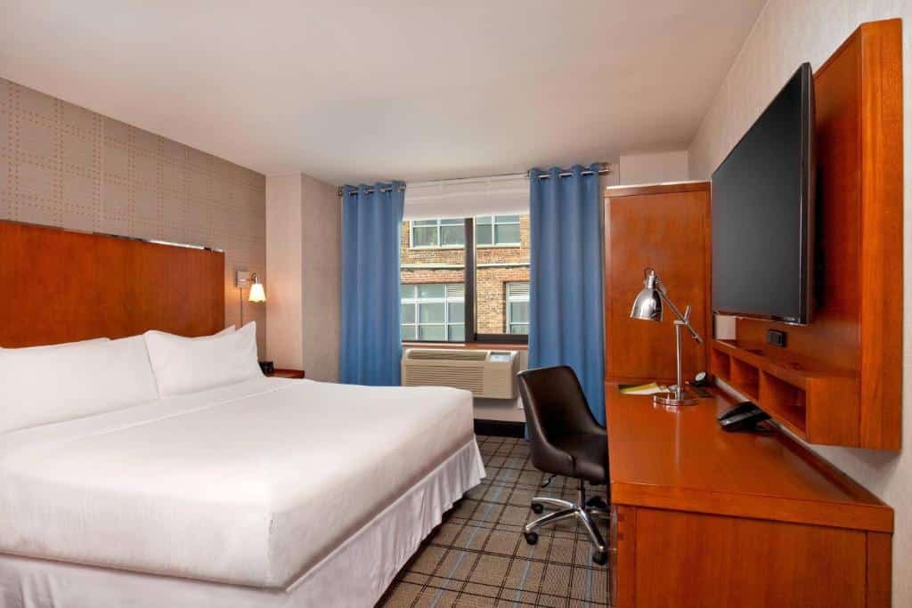 suíte do Four Points by Sheraton Midtown - Times Square com uma cama de casal à esquerda, em frente a um painel de madeira com televisão presa à parede e uma mesa abaixo, acompanhada de uma cadeira estofada e giratória.
