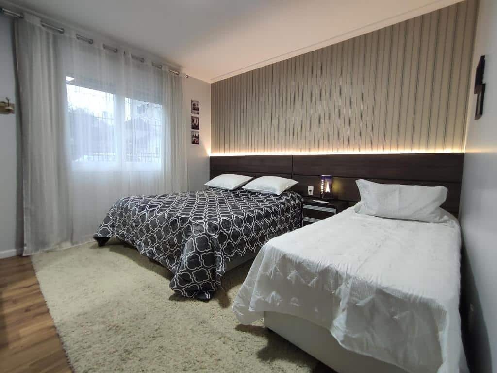 Quarto em Gramado Garden Centro, uma cama de casal e uma de solteiro, iluminação indireta, uma sacada com cortina e um tapete