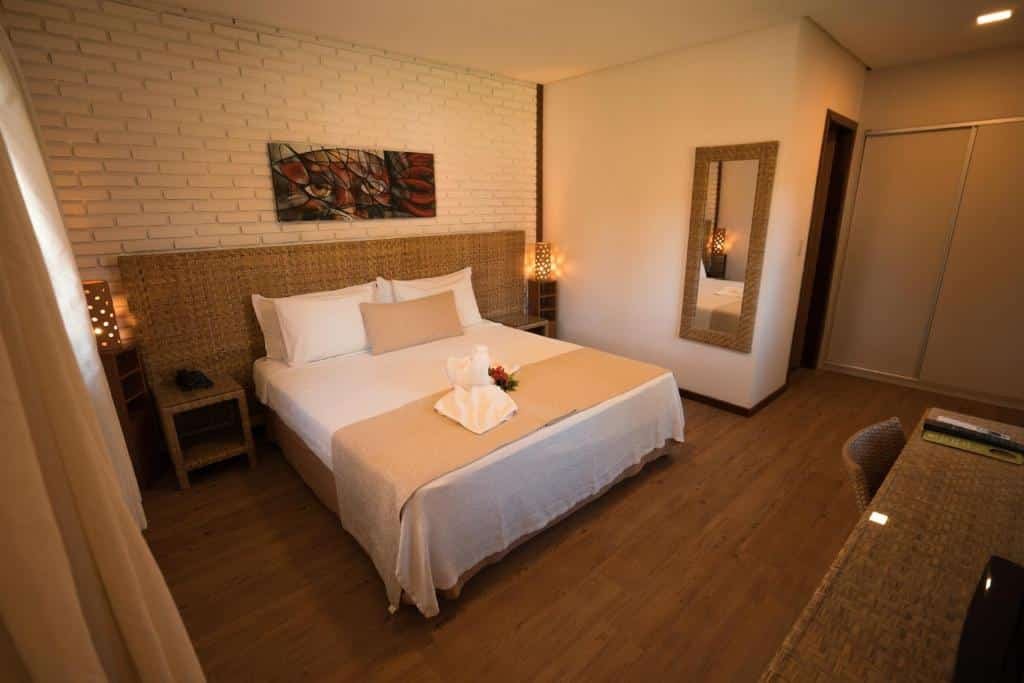 suíte do Grand Oca Maragogi Resort, com uma cama de casal com travesseiros em cima e uma cabeceira rústica feita de material artesanal. Há um quadro acima da cama com desenho abstrato e um espelho na parede à direita. É possível ver prte de uma mesa comuma cadeira e uma porta dando acesso ao banheiro.