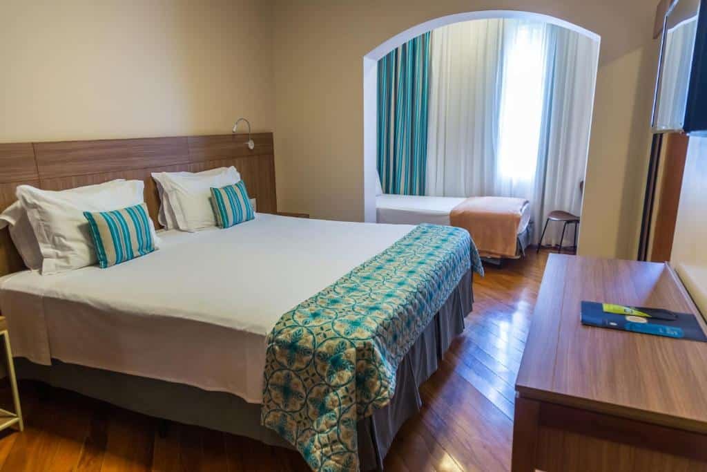 Quarto no Grand Resort Serra Negra com uma cama de casal no primeiro ambiente e uma televisão, no ambiente ao lado uma cama de solteiro e uma cadeira, além de uma janela com cortinas