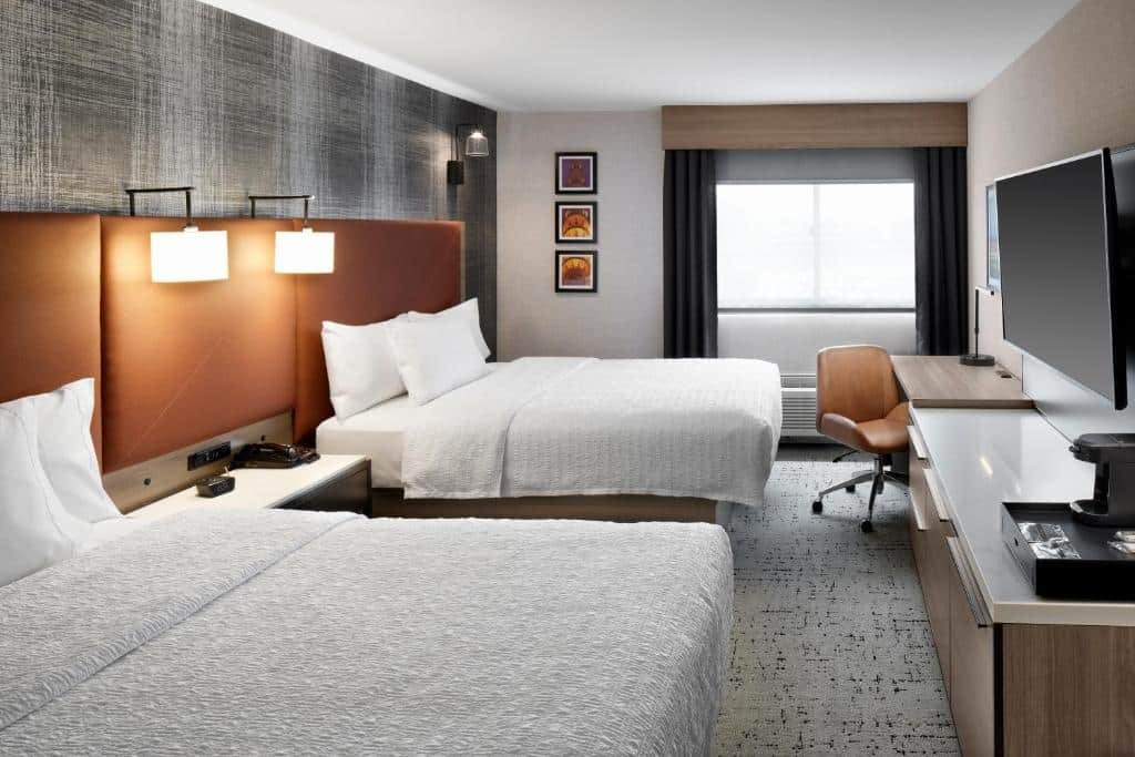 Imagem de um quarto acolhedor e funcional no Hampton Inn Newark Airport. O quarto apresenta uma decoração contemporânea com cores suaves e móveis práticos. A cama é espaçosa e confortável, com lençóis macios e bem arrumados. Nas laterais da cama, há mesas de cabeceira com luminárias de leitura. Ao lado direito, há uma escrivaninha bem iluminada com uma cadeira ergonômica para trabalho ou estudo. Na parede oposta à cama, há uma TV de tela plana para entretenimento. O quarto também inclui um armário para guardar pertences pessoais. A atmosfera do quarto é agradável e convidativa, proporcionando conforto aos hóspedes. O Hampton Inn Newark Airport é um hotel localizado perto do aeroporto de Newark, oferecendo praticidade para viajantes.
