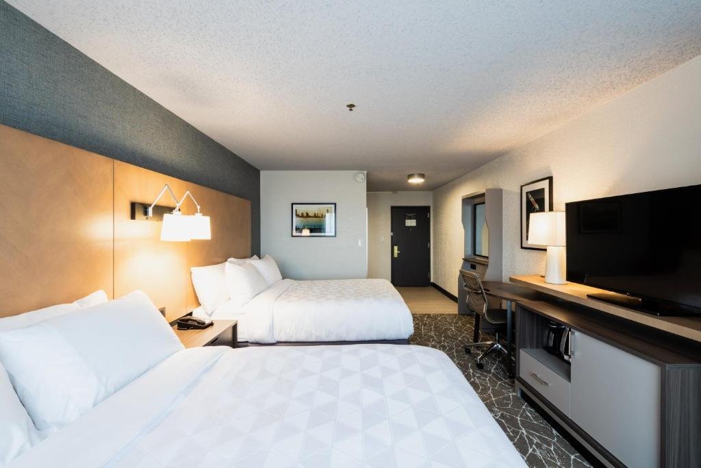 Imagem de um quarto no Holiday Inn Newark International Airport. No centro do quarto, há uma cama king-size com lençóis brancos e travesseiros macios. À direita da cama, há uma mesa de cabeceira com uma luminária de leitura. Na parede à esquerda da cama, há uma TV de tela plana montada. Ao lado direito da imagem, há uma área de trabalho com uma escrivaninha e uma cadeira. O quarto é iluminado de forma suave e possui uma atmosfera aconchegante. O Holiday Inn Newark International Airport é um dos hotéis perto do aeroporto de Newark.