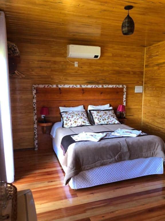 quarto da Hospedagem Família Cruz um dos chalés em Urubici, com cama de casal ao centro, mesinha e luminária de ambos os lados, ar-condicionado acima, de um lado é possível ver uma grande janela com cortinas, as paredes são de madeira.