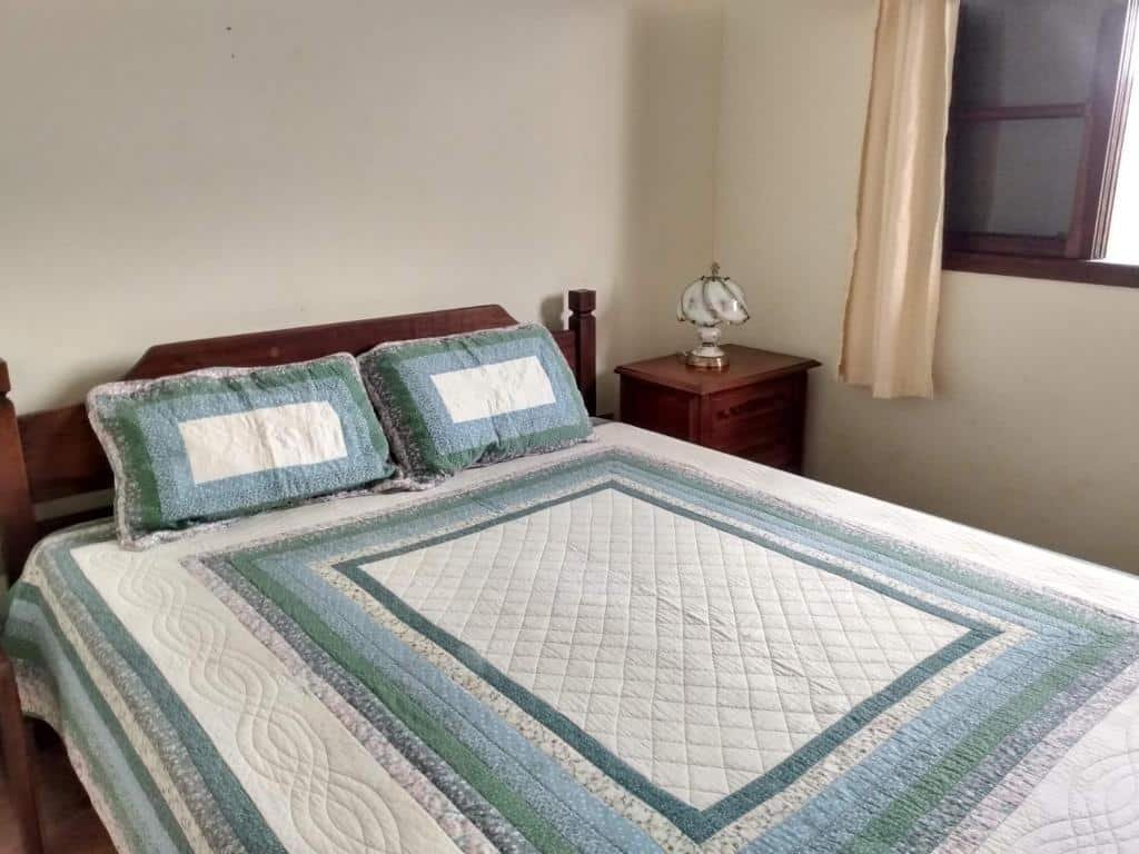 Quarto da Hospedaria Namastê. Uma cama de casal com dois travesseiros. No lado direito da cama uma cômoda e a janela do quarto.