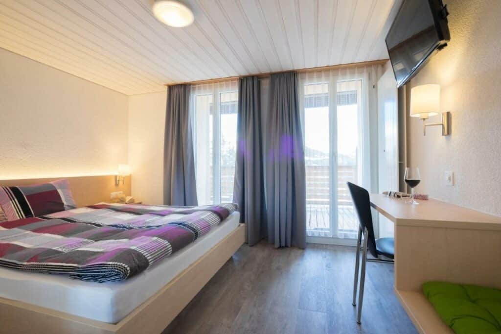Quarto do Hotel Alpenblick Mürren com cama de casal do lado esquerdo da imagem em frente a cama uma mesa com cadeira e em cima a TV presa na parede. Representa hotéis em Lauterbrunnen.
