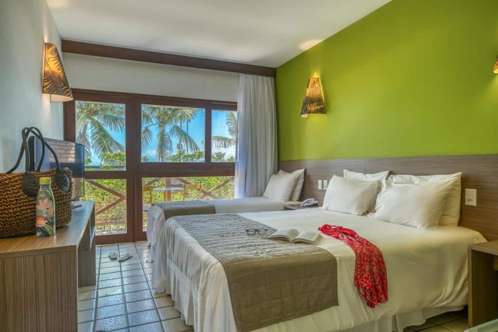 quarto do Hotel Areias Belas, em Maragogi, com uma cama de casal e outra de solteiro e uma varanda com vista de árvores e uma mesa com duas cadeiras