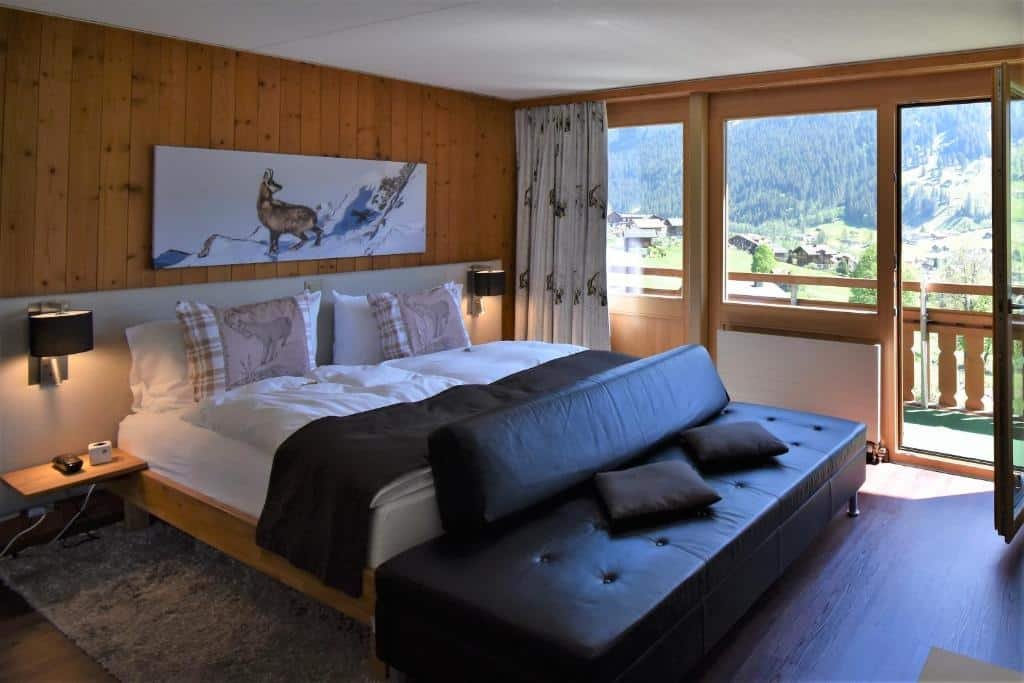 Quarto do Hotel Cabana com cama de casal do lado esquerdo da imagem no centro do quarto com um sofá no pé da cama e do lado esquerdo da cama uma porta de vidro com acesso a varanda. Representa hotéis em Lauterbrunnen.
