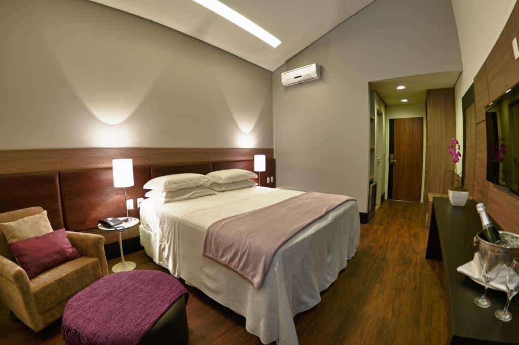 Quarto no Cordilheira Hotel com uma cama de casal, uma cabeceira com iluminação indireta, duas poltronas e uma mesinha, uma televisão e um ar-condicionado, para representar hotéis em Serra Negra