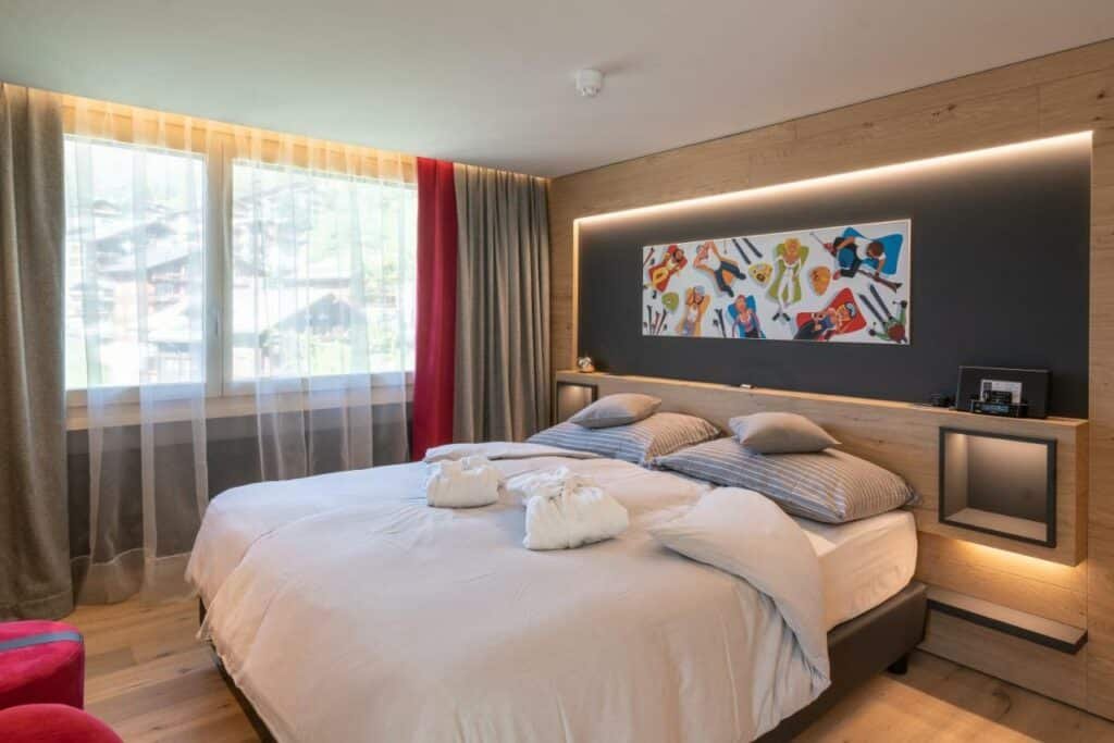 Quarto do Hotel Eden Wellness com cama de casal do lado direito da imagem do lado direito da cama uma janela ampla com cortinas e em frente a cama dois puffs. Representa onde ficar em Zermatt.