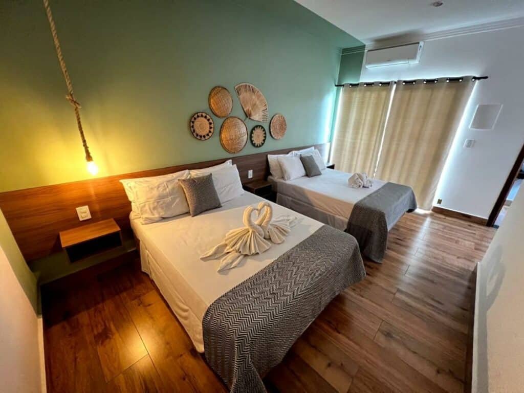 Quarto no Hotel Fazenda Vale da Cachoeira com uma cama de casal, uma cama de solteiro, uma cabeceira, um ar-condicionado, uma varanda com cortinas, o chão é de madeira e a decoração é rústica, para representar hotéis em Serra Negra