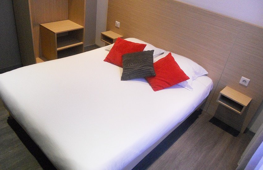 Quarto do Hôtel de France. Uma cama de casal com uma prateleira de apoio e tomada de cada lado.