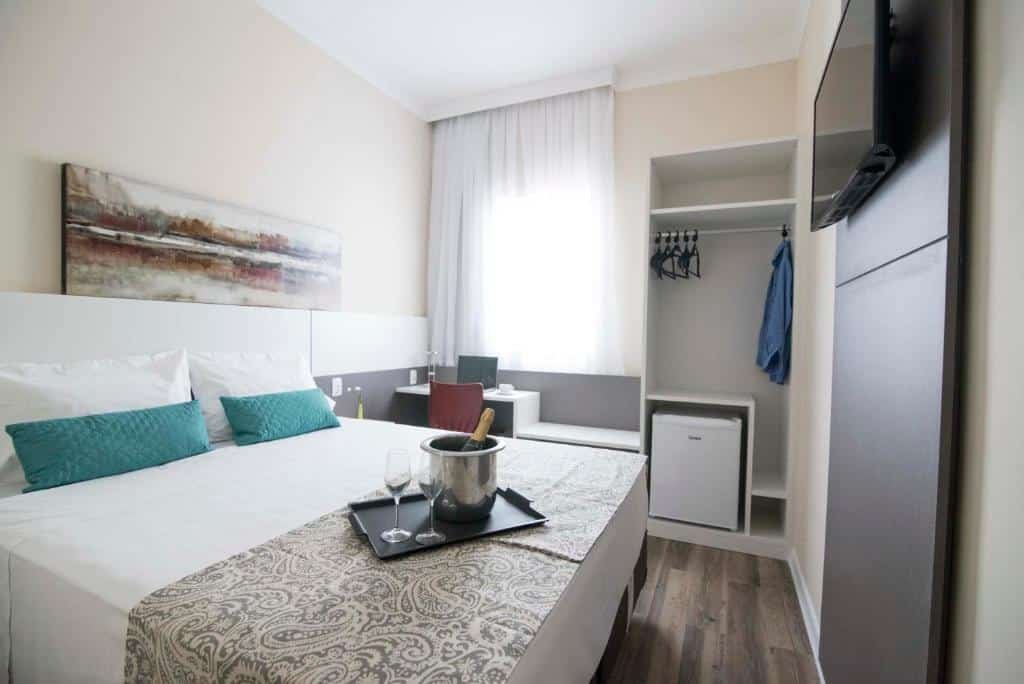 suíte do Hotel H2 Congonhas com uma cama de casal à esquerda, uma bandeja com duas taças de vidro e um balde com champanhe em cima dos lençóis, e ao fundo do quarto uma pequena janela de canto coberta por cortinas brancas transparentes, uma pequena mesa branca com uma cadeira vermelha abaixo, e um armário branco e sem porta ao seu lado.