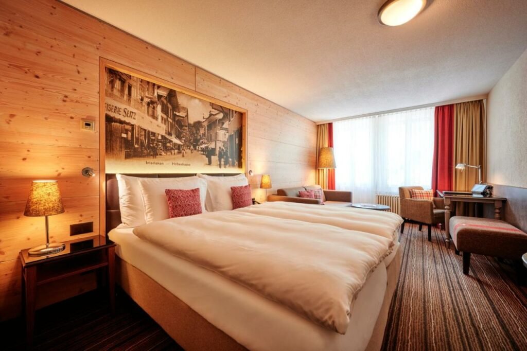 Quarto do Hotel Interlaken com cama de casal do lado esquerdo da imagem uma cômoda em cada lado com luminária e do lado esquerdo da cama um sofá e em frente ao sofá uma mesa de trabalho com poltrona. Representa hotéis em Lauterbrunnen.