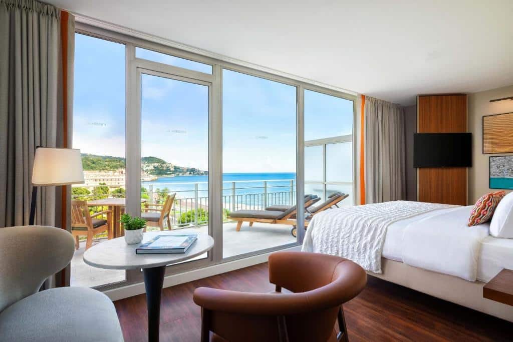 Quarto do Le Meridien Nice. A cama está no lado direito do quarto com uma TV a frente pendurada na parede. No lado esquerdo há um abajour de chão, uma mesa de centro e duas cadeiras. A parede da frente do quarto é toda de vidro e dá acesso a uma enorme varanda com vista para o mar, uma mesa, cadeiras e espreguiçadeiras. Imagem para ilustrar o post hotéis em Nice.