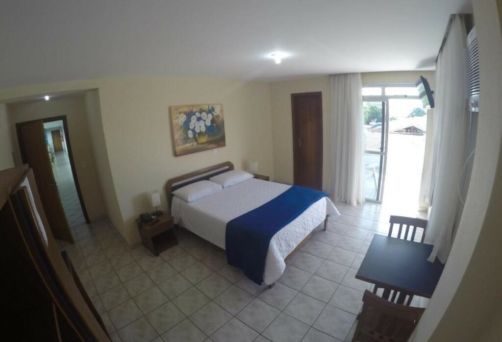 Quarto do Hotel Marlin Azul. Uma mesa de madeira com duas cadeiras no lado direito. Atrás, uma televisão e a porta com acesso a sacada. No lado esquerdo, uma cama de casal e atrás a porta do banheiro. No canto do lado esquerdo a porta do quarto e um guarda-roupa.