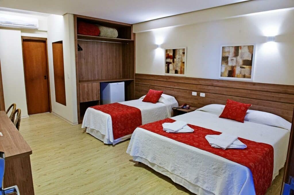 Quarto com duas camas, uma de casal e outra de solteiro. Ao lado há um armário e um mini frigobar. No canto da imagem é possível ver uma mesa com cadeira. Foto para ilustrar post sobre hotéis em Campo Grande.