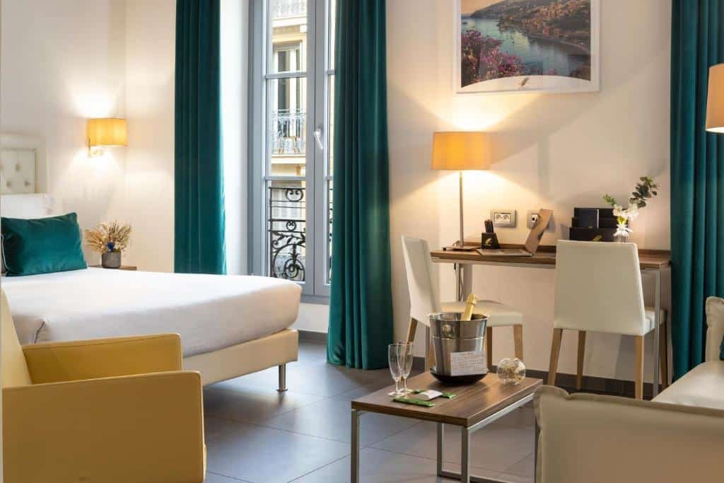 Quarto do Hôtel Nice Azur Riviera. A cama está encostada na parede do lado esquerdo do quarto, em frente há uma mesa de centro com um balde de gelo com champanhe e atrás há um sofá. Na parede direita há uma porta de vidro que dá acesso a varanda e uma mesa com duas cadeiras. Imagem para ilustrar o post hotéis em Nice.
