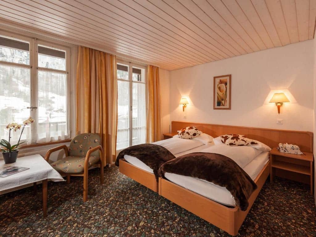 Quarto do Hotel Oberland com cama de casal no centro do quarto com duas cômodas em cada lado da cama e do lado esquerdo da cama uma cadeira com uma mesa. Representa hotéis em Lauterbrunnen.