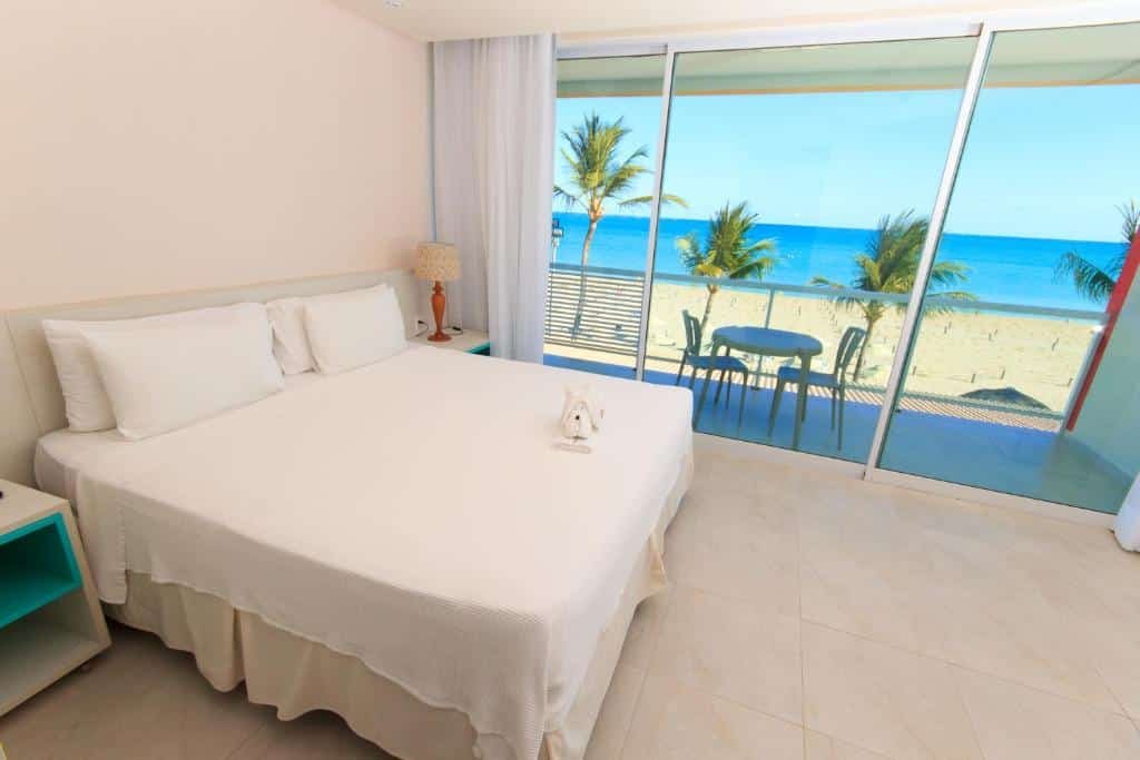 quarto do Hotel Praia Dourada, com uma cama de casal, e uma varanda à direta com porta de vidro, vista para o mar e uma mesinha redonda com duas cadeiras