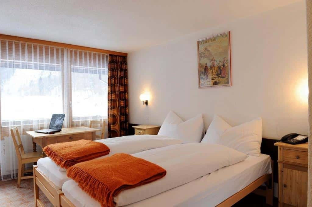 Imagem do quarto do Hotel Schützen Lauterbrunnen com cama de casal do lado direito da imagem e uma cômoda em cada lado da cama do lado direito da imagem uma mesa quadrada com duas cadeiras. Representa hotéis em Lauterbrunnen.