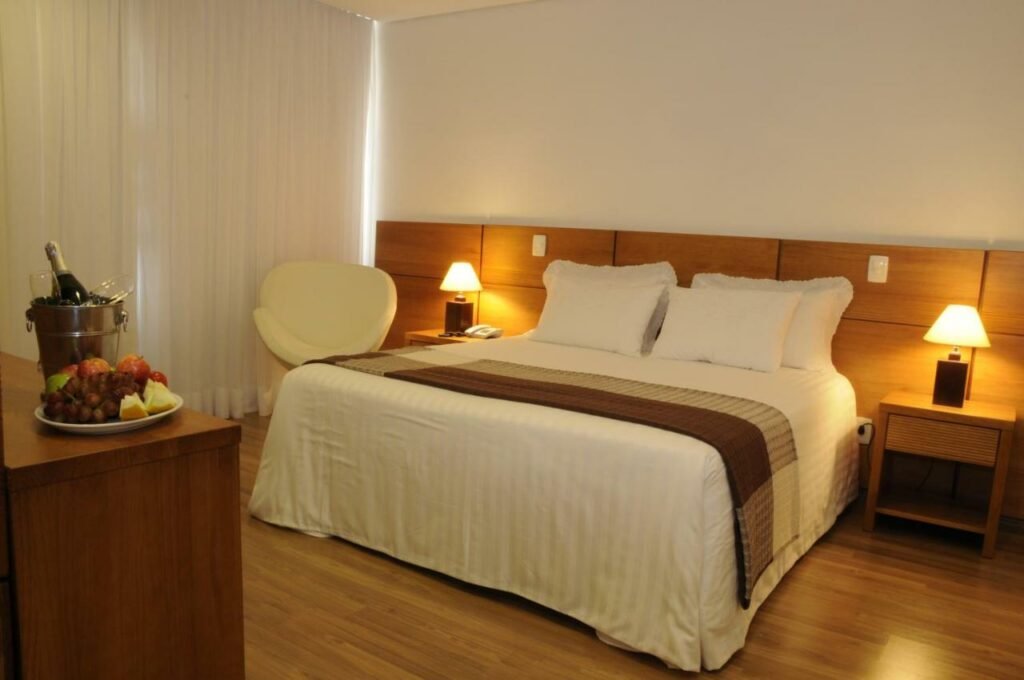 Quarto do Hotel Shelton com uma cama de casal, uma janela com cortinas, uma poltrona, uma cômoda com taças, vinho e frutas sob ela, o chão é de madeira, e há duas mesinhas de cabeceira com abajures dos dois lados da cama, para representar hotéis em Serra Negra