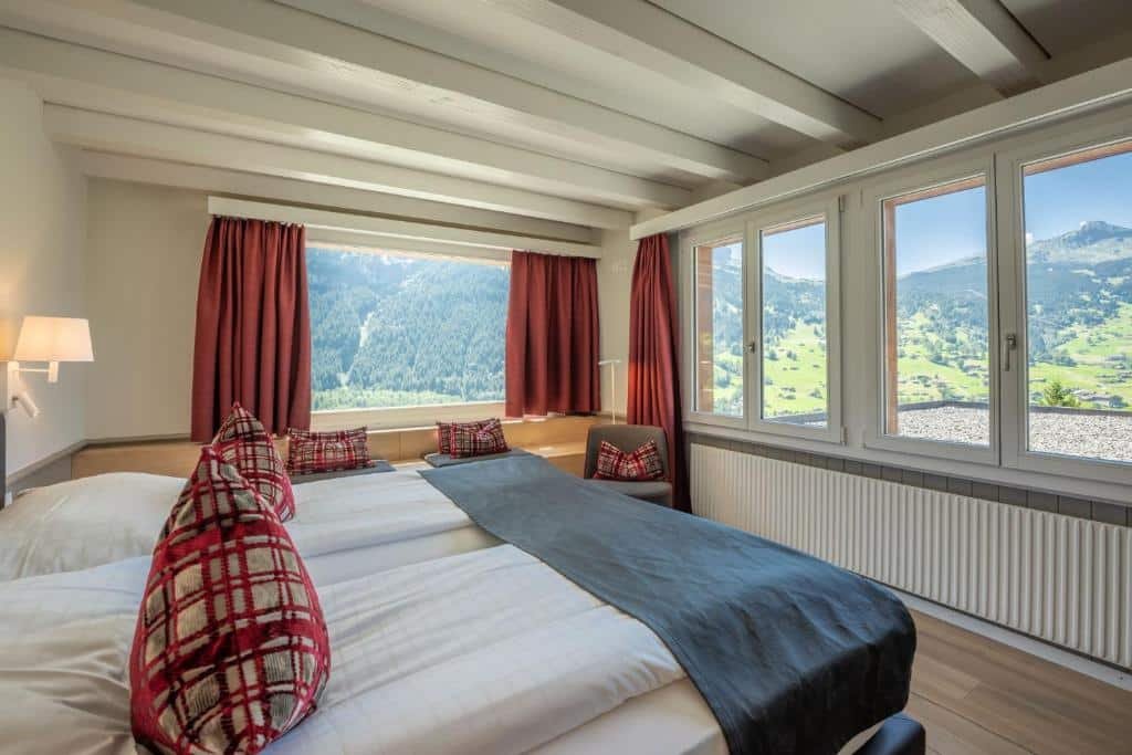 Quarto do Hotel Spinne Grindelwald com cama de casal do lado esquerdo da imagem em frente a cama há várias janelas com vista para as montanhas. Representa hotéis em Lauterbrunnen.