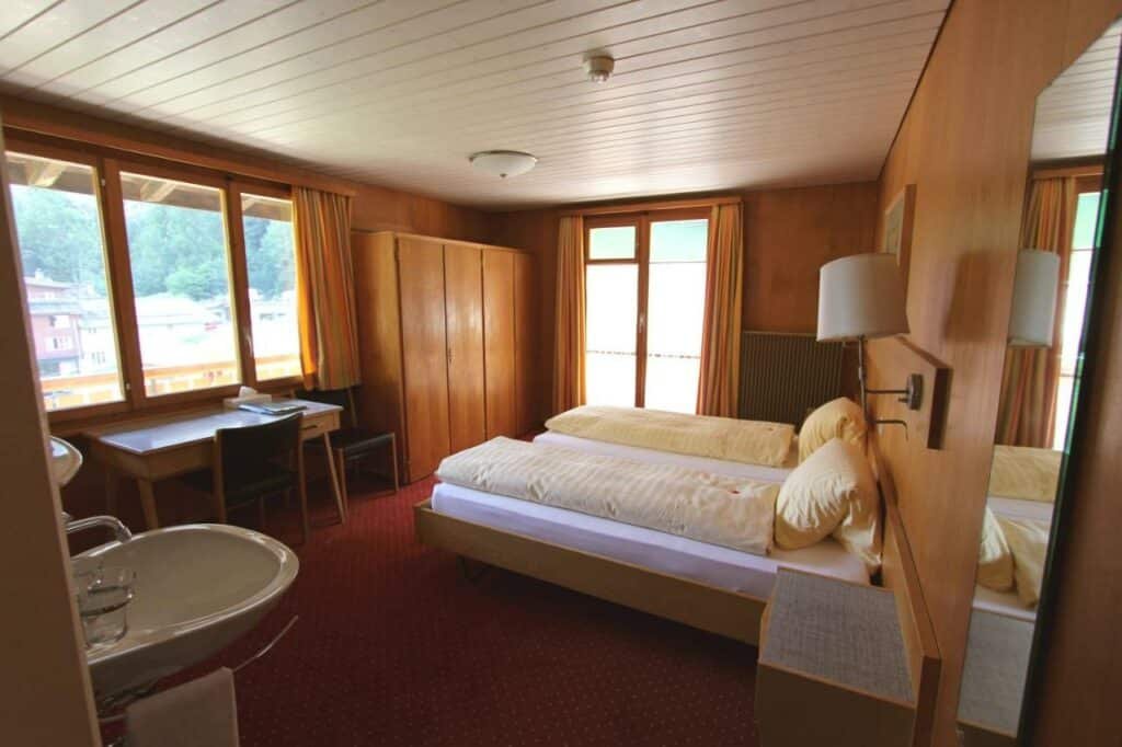 Quarto do Hotel Staubbach com cama de casal do lado direito da imagem no centro do quarto em frente a cama uma mesa com cadeira e do lado direito da mesa um guarda-roupa. Representa hotéis em Lauterbrunnen.