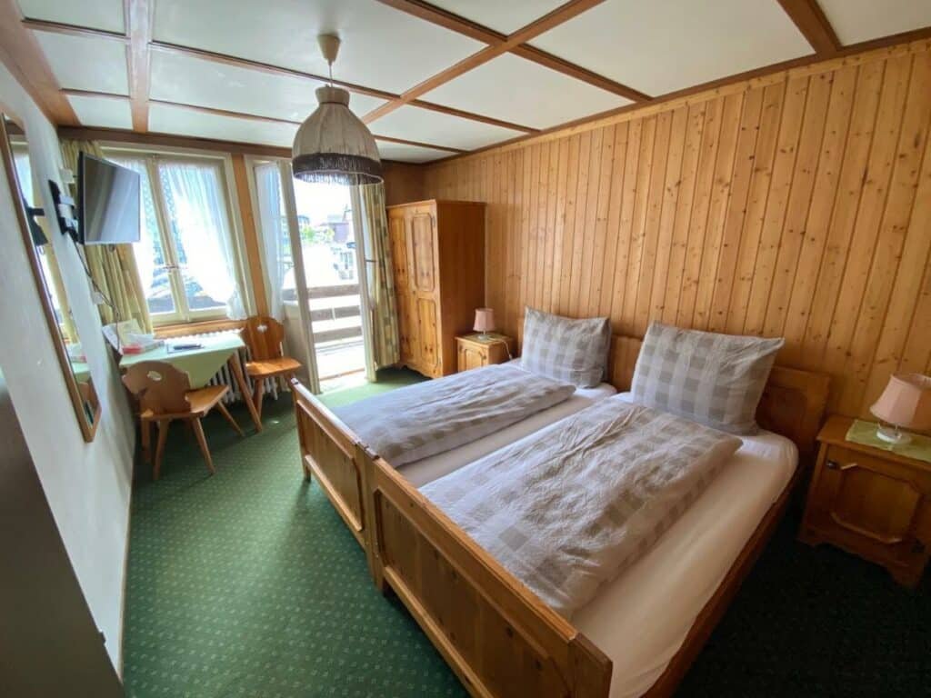 Quarto do Hotel Steinbock com cama de casal do lado direito da imagem com uma cômoda em cada lado da cama com luminária do lado direito da cama um guarda-roupa e uma mesa com duas cadeiras. Representa hotéis em Lauterbrunnen.