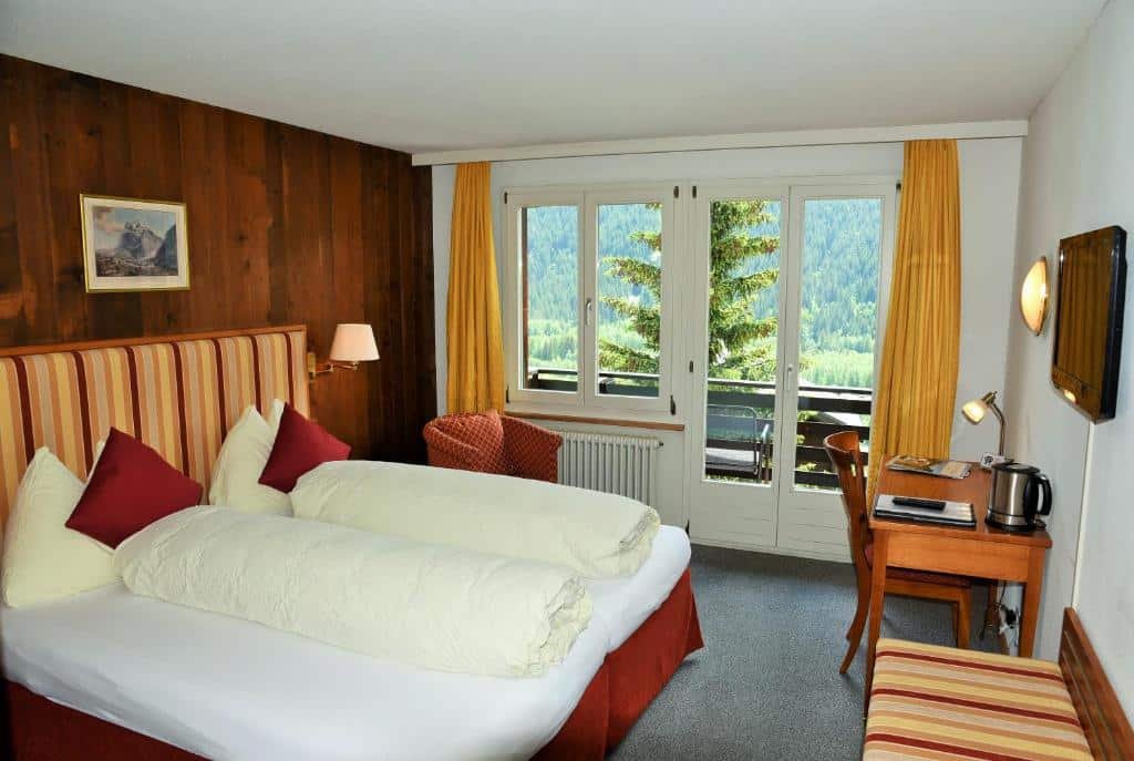 Quarto do Hotel Tschuggen com cama de casal do lado esquerdo da imagem a frente e do lado esquerdo da cama uma poltrona e em frente a poltrona uma cômoda com TV.