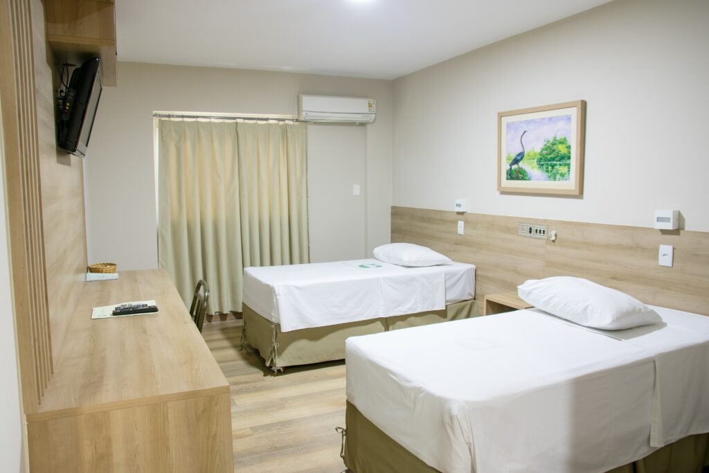 Quarto com duas camas, uma mesa com cadeira e televisão acima. No canto é possível ver uma janela com cortina e um ar-condicionado.