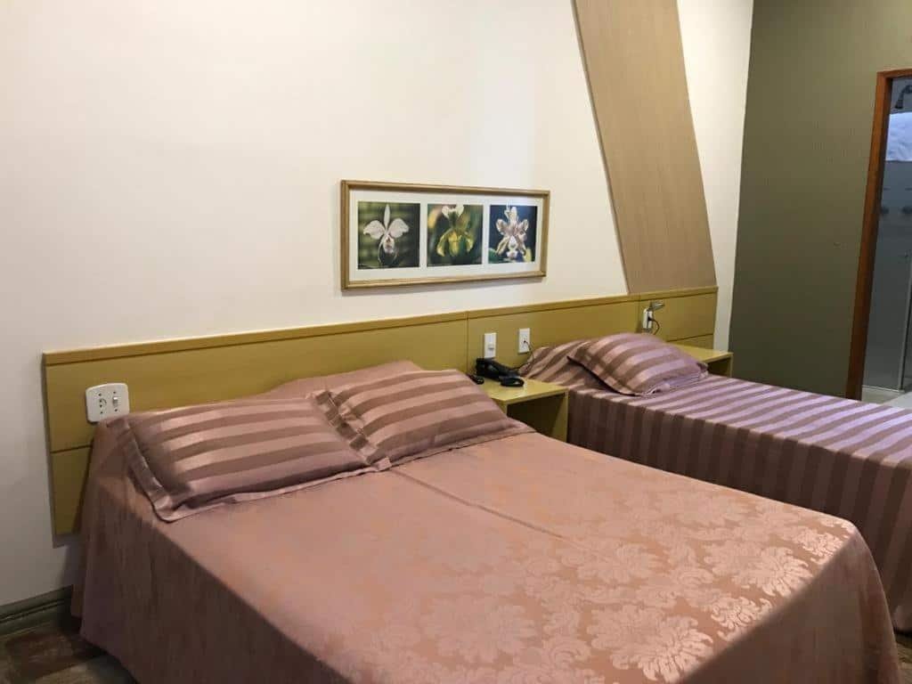 quarto do Hotel Verdes Mares em Ouro Branco com uma cama de casal e uma de solteiro, lado a lado, separados apenas por uma pequena cômoda no meio, onde há um telefone preto. Acima das cabeceiras há um quadro estreito com três fotos de flores distintas