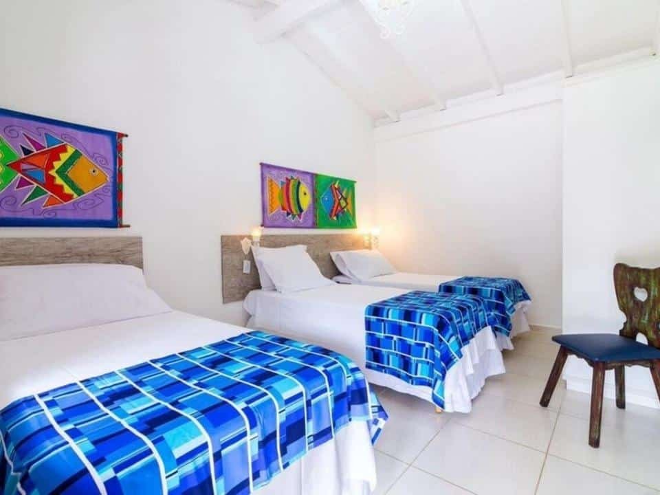 Quarto no Hotel Village Enseada, uma cama de casal e duas de solteiro, tudo em tons de branco e azul, uma cadeira rústica, alguns quadros com peixes desenhados, para representar pousadas na Praia da Enseada em Ubatuba