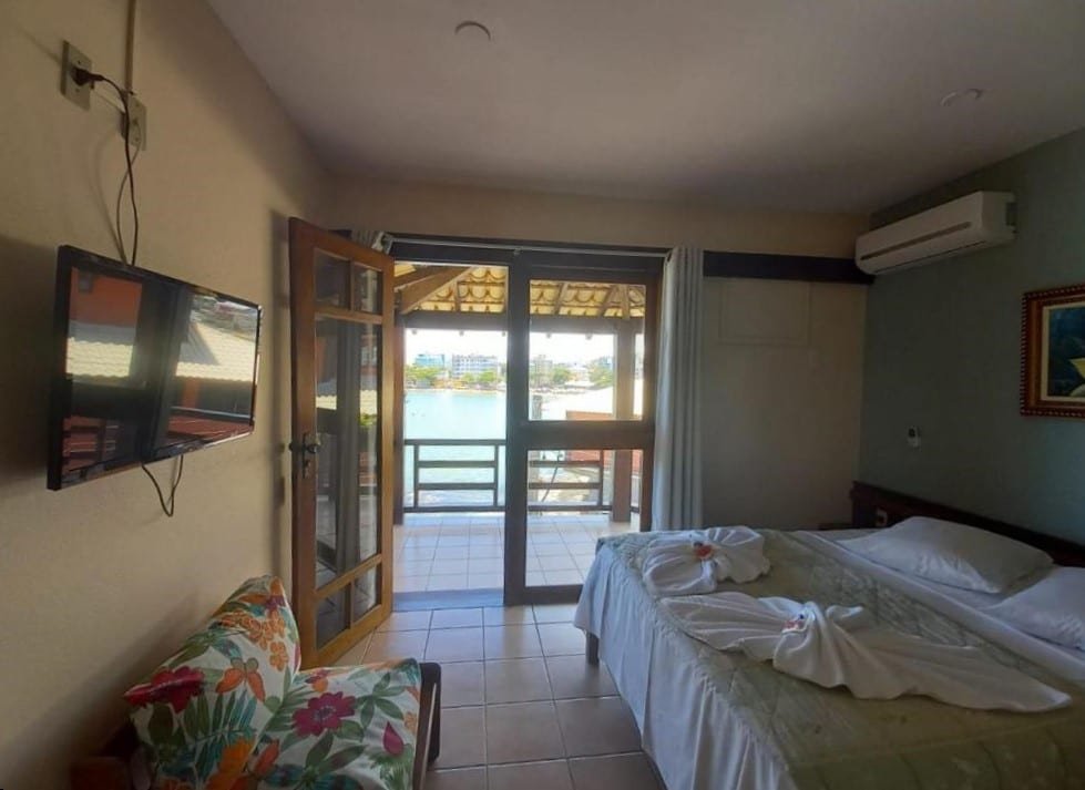 Quarto do Hotel Pontal das Rochas. Uma cama de casal no lado direito com um ar-condicionado em cima. De frente, um sofá e uma televisão. No fundo, a porta de vidro com acesso para a sacada e vista para o mar. Foto para ilustrar post de pousadas em Iriri (ES).
