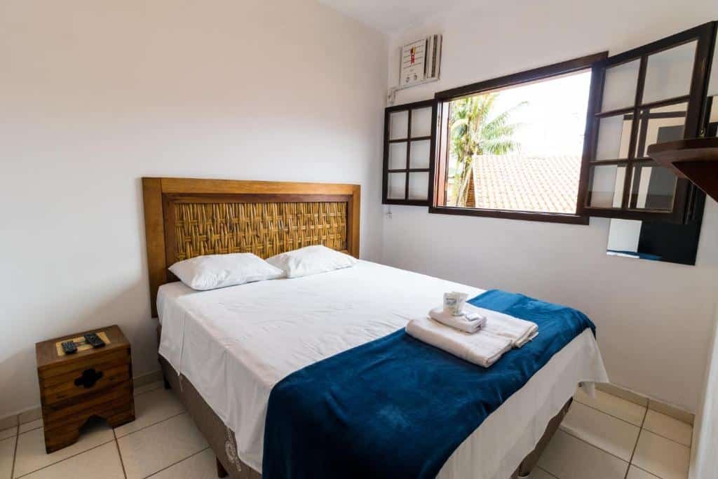 Quarto na HOTELARE Bóra Morá Pousada, uma cama de casal, com travesseiros e toalhas sob a mesma, uma cabeceira e uma mesinha de madeira rústica e uma ampla janela, para pousadas na Praia do Tenório