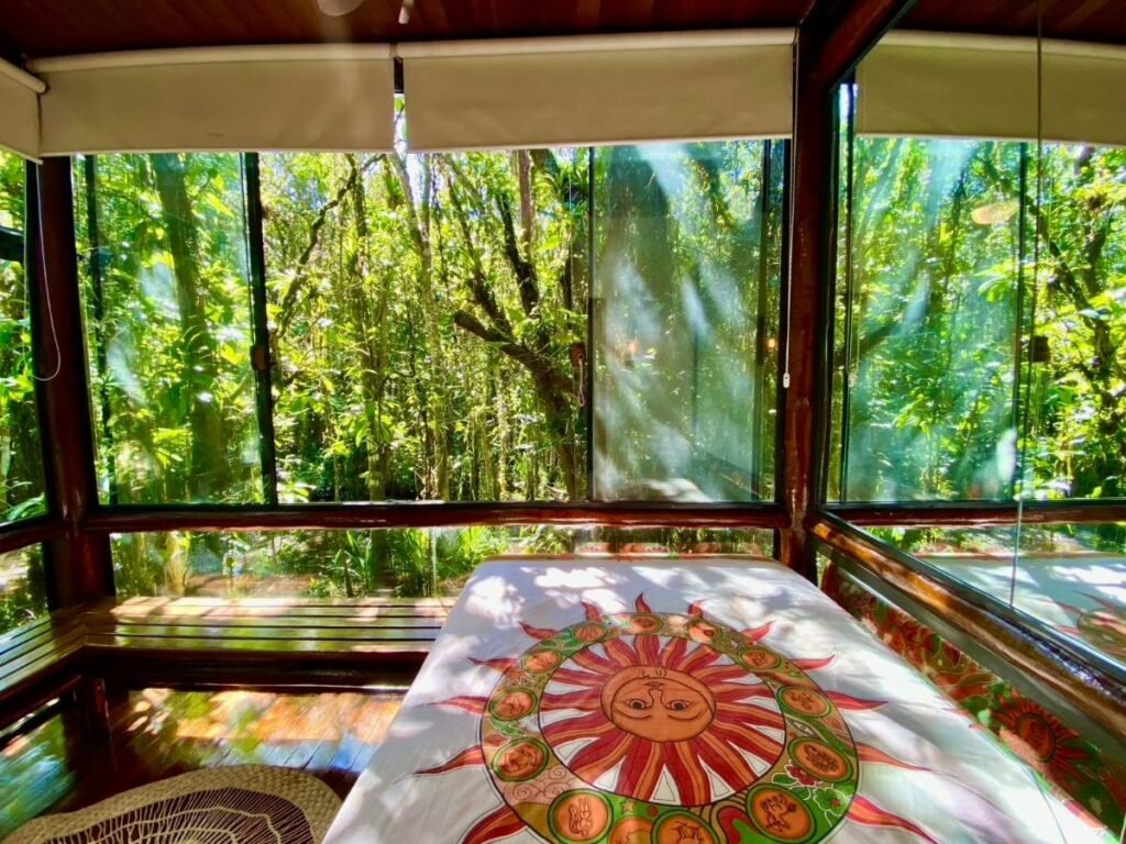 Quarto do Housing 31 - Cond. Mata Azul com cama de casal do lado direto da imagem com uma colcha de sol em cima da cama, o quarto fica em volta de paredes de vidro que dá para vê a floresta em volta.